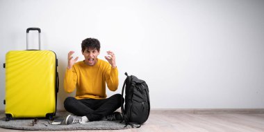 Stresli genç gezgin, beyaz arka planda hayal kırıklığı içinde bavuluyla oturup bağırıyor. Pankart