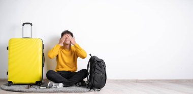 Genç gezgin gözlerini elleriyle kapatarak beyaz arka planda izole edilmiş bir bavulla oturuyor. Pankart