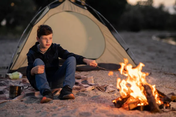Teen boy roasts marshmallows on a campfire in the evening. Copy space