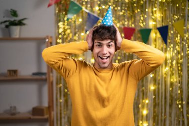 Neşeli şapkalı komik bir adamın yakın plan fotoğrafı olumlu haberleri dinleyin kollarınızı başınızın üstüne koyun ve günlük sarı süveter giyin. evde doğum günü partisi konsepti
