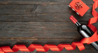 Sevgililer Günü tebrik kartı hediye kutusu ve ahşap arka planda saten kurdeleli kırmızı şarap şişesi. Tebrik etmek için en üstteki dairede yer var. Boşluğu kopyala