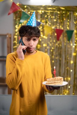 Parti şapkalı bir adam doğum gününü evde kutluyor cep telefonuyla konuşuyor doğum günü pastasını tutarken kötü bir tebrik alıyor. Boşluğu kopyala
