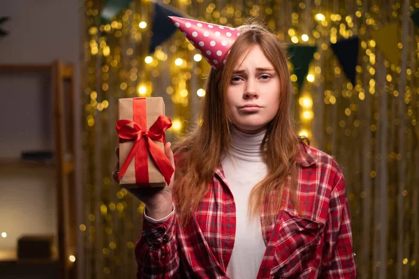 Picture of upset frustrated young woman holding present box with ribbon at party at home, being in bad mood because of problems at work, lonely birthday. Human emotions and feelings. Copy space