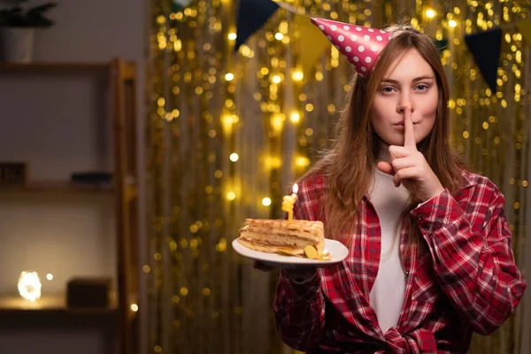 Girl in a birthday hat holding a cake and covering her mouth with a finger having a secret Celebrating birthday at home. Copy space