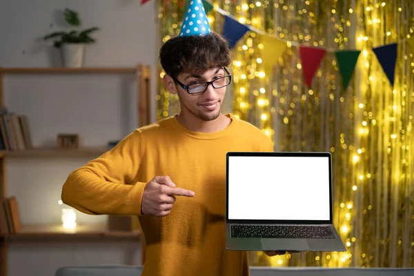 Joyful arabian or indian guy in party hat, freelancer or student, holding an open laptop with empty mock-up space and points finger on it, celebrate his birthday at home. Copy space