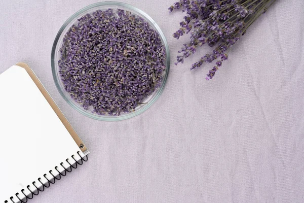 Lavender flowers in glass bowl, notebook for recipes and branches on light background, toned. Invitation, spa, recipe concept. Top view, flat lay, copy space, layout design