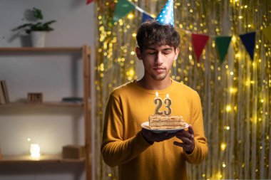 Adam 23. doğum gününü evde kutluyor, elinde mumlarla pasta tutuyor ve dilek tutuyor.