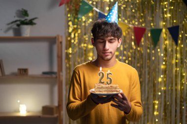 25. yaş gününü evde kutlayan adam elinde mumlarla pasta tutuyor ve dilek tutuyor.