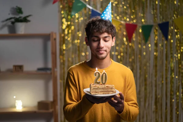 20th birthday. Twenty year old guy celebrates his holiday at home, blows out candles on a birthday cake. Man in party hat make a wish. Copy space
