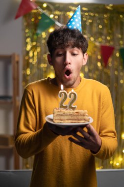 Tatiller ve kutlamalar. Heyecanlı genç adam 22. yaş gününü kutluyor, pastaya mum üflüyor, parti şapkası takıyor ve eğleniyor, ev dekorasyonunun başında duruyor. Boşluğu kopyala