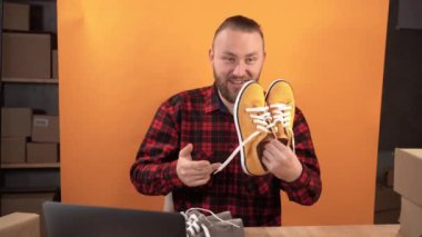 Genç bir işletme sahibi bir video stüdyosunda oturmuş bir online mağaza için mal değerlendirmesi yapıyor. Kameraya bakar sarı ve gri spor ayakkabıları karşılaştırır. İş dünyası sosyal medya etkileyiciName.