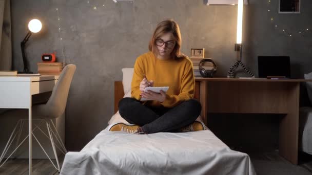 一个女学生穿着橙色毛衣坐在大学宿舍的床上 手里拿着笔记本 用笔写字 青少年做作业的概念 复制空间 — 图库视频影像