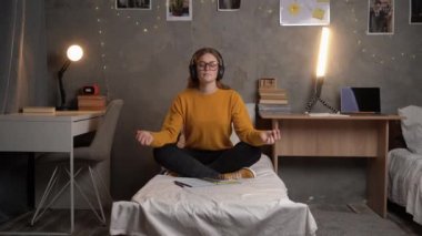A student girl sits in a college dormitory on a bed in a lotus position in an orange sweater with glasses, listens to music on headphones and meditates. copy space.