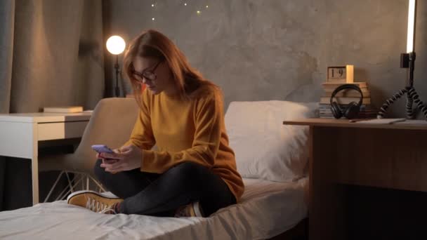 学生の女の子はオレンジのセーターを着たベッドの上の大学の寮に座り スマートフォンを手に持ち 指でメッセージを打ちます ソーシャルメディアです コピースペース — ストック動画