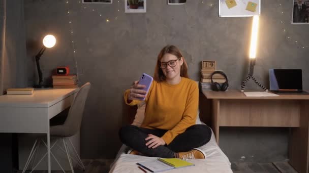一名女生穿着橙色毛衣坐在大学宿舍的床上 戴着眼镜 想用智能手机在网上打一个视频电话 复制空间 — 图库视频影像