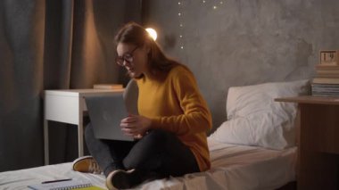 a girl student in glasses sits on bed in college or university dormitory dressed in orange sweater. A teenager is looking for information online in a laptop and making notes in a notebook. copy space