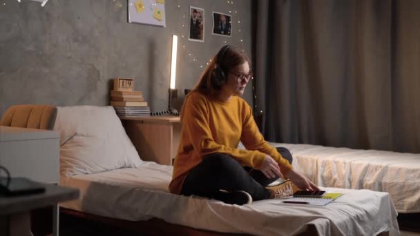 学生の女の子は 眼鏡をかけたオレンジ色のセーターを着た蓮の花の位置のベッドの上の大学の寮に座って ヘッドフォンで音楽を聞き 瞑想します コピースペース — ストック動画