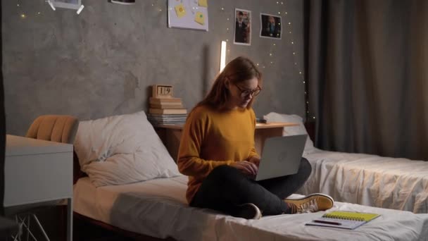 眼鏡をかけた若い女子学生の肖像画がラップトップで働いているオレンジのセーターを着た大学の寮のベッドに座っている コピースペース — ストック動画