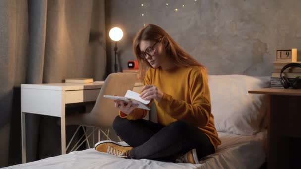 女子大生はオレンジのセーターを着たベッドの上の大学の寮に座りノートを手にノートを持ってノートを探しながら並べ替えます 十代の若者が宿題をするという概念 コピースペース — ストック動画