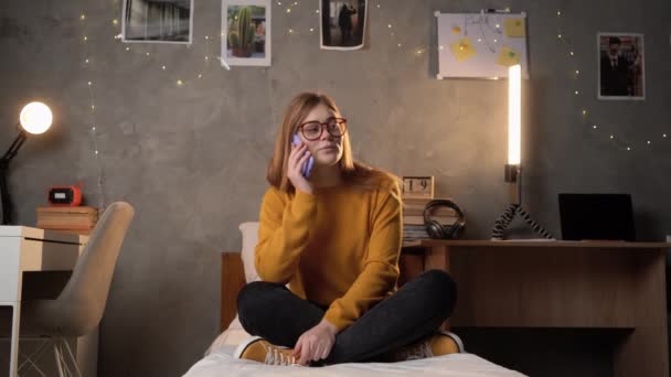 一个女学生穿着橙色毛衣坐在大学宿舍的床上 用智能手机打电话 复制空间 — 图库视频影像