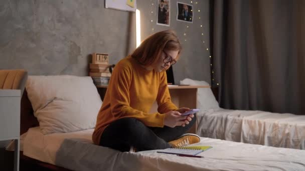一位女学生穿着橙色毛衣坐在大学宿舍的床上 戴着眼镜 用智能手机在网上搜寻信息 并在笔记本上记笔记 复制空间 — 图库视频影像
