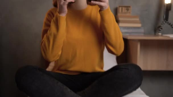 学生の女の子は 眼鏡をかけたオレンジのセーターを着たベッドの上の大学の寮に座って ワイヤレスヘッドフォンをして 彼女のスマートフォンで音楽をオンにし 耳を傾けます コピースペース — ストック動画
