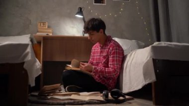a young Arab student sits at night on the floor in a hostel among open paper books looking for the necessary information. The concept of preparing a homemade creative task. copy space.