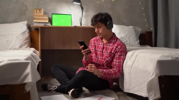 一个年轻的阿拉伯学生晚上坐在大学宿舍的地板上 用无线耳机听音乐 手里拿着智能手机打字 打印出一条信息 — 图库视频影像