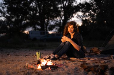 Woman with coffee warming near campfire in forest listening music in headphones. Copy space