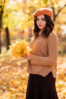Beautiful happy young woman in park in fall holding leaves. Teenage girl standing outdoors in autumn. Vibrant colors. Copy space