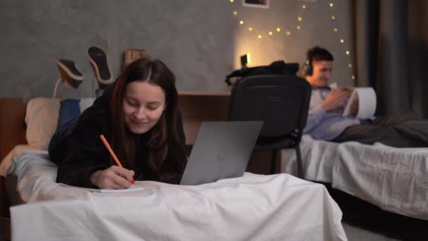 年轻的女学生晚上躺在大学宿舍的床上 用她的笔记本电脑用钢笔在笔记本上记录网上信息 复制空间 — 图库视频影像