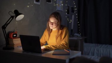 girl teenager student sits at night at a table in a college dormitory looking for information online using a laptop. copy space.