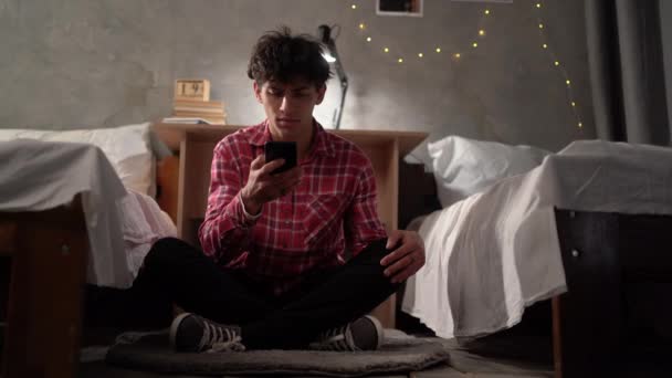一个在大学宿舍里的年轻学生坐在地板上 看着他的智能手机 举着他的头 压力和沮丧 青少年中的欺侮和勒索 复制空间 — 图库视频影像