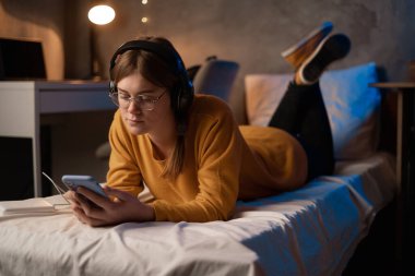 Girl using mobile phone lying on bed in dorm, female student listening to music with headphones in dormitory room. Copy space