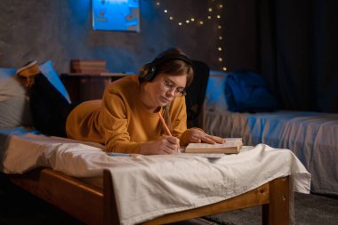 Female college student reading thick book and taking notes in textbook on bed. Copy space