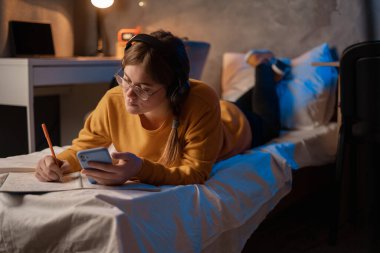 Studying at dormitory online. Young woman studying or prepare for exam using cellphone while lying on the bed at night time. Copy space