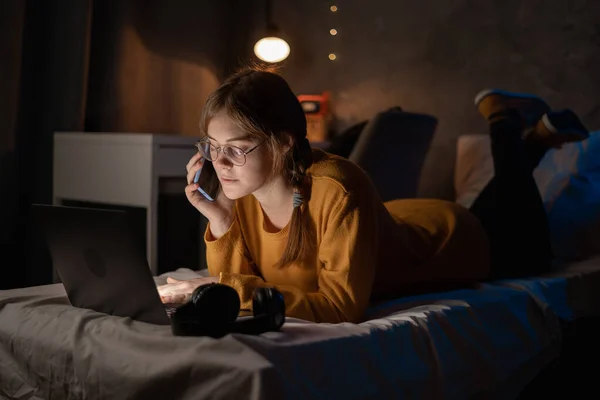 Female college student talking on mobile phone, lying on bed working on laptop at night in dormitory. Copy space