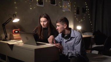 two teenagers are sitting at a table at night in a hostel, preparing a creative task and intensively discussing something. Searching for information on a laptop