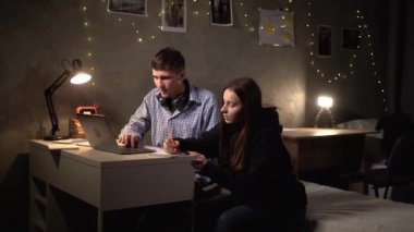 two students, a guy and a girl, sit at a table at night in a dorm room and prepare together for a creative task using a laptop. Make notes in a notebook with a pen. copy space.