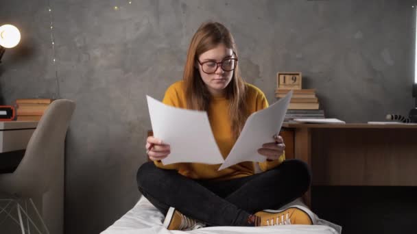 戴眼镜的少女坐在大学宿舍的床上 手里拿着一张纸看书 准备考试的概念 复制空间 — 图库视频影像