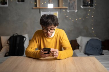 Attractive arab student man sitting at table studying holding mobile phone surfing internet received message from friend chatting about weekend plans in dormitory room. Copy space