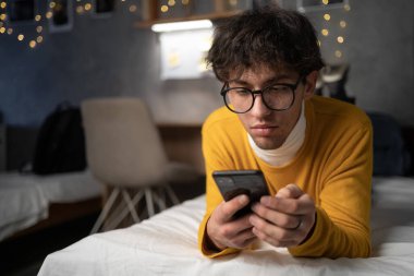 Young man studies while lying on the bed using smartphone in the evening in student dormitory. Copy space