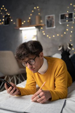 Male college student lies on bed in shared house working on smartphone and writing notes in notebook. Copy space