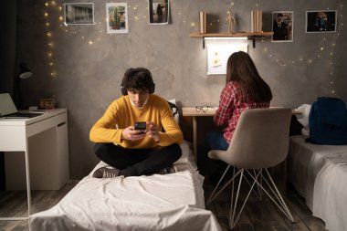 Portrait of a guy student sitting on the bed in the evening in a college dormitory and chatting on cellphone. University lifestyle concept. Copy space.