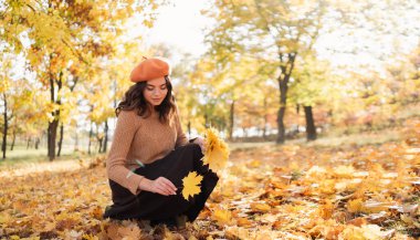 Happy smiling young woman in park on sunny autumn day collect maple leaves. Copy space