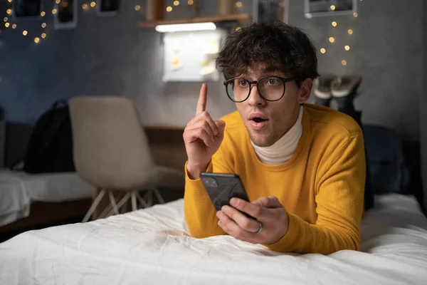 Teenager boy using smartphone on the bed surprised with an idea or question pointing finger with happy face, number one. Copy space