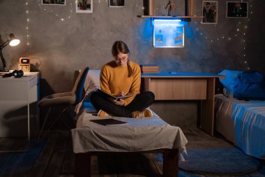 Female college student sitting on bed doing homework using copybooks and making notes preparing for exam at night in dormitory room. Copy space
