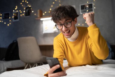 Excited happy young man winning feeling joy using smartphone winning lottery game, betting, getting cashback online gift using smartphone lying on bed in dormitory. Copy space