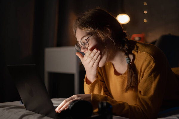 Tired young woman yawning, lying on bed with laptop, lazy student doing homework, sleepy girl working on computer in dormitory, sleepless night, lack of sleep and boredom concept