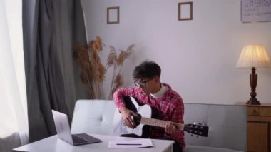 Man with acoustic guitar sitting on sofa with laptop online music lessons. Arabic young guitar player practicing musical instrument. Copy space